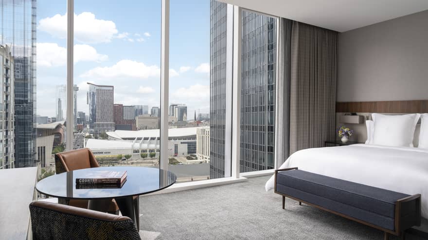 Hotel room with a king-size bed and a small round table with two leather chairs, featuring floor-to-ceiling windows offering a cityscape view, including the Bridgestone Arena