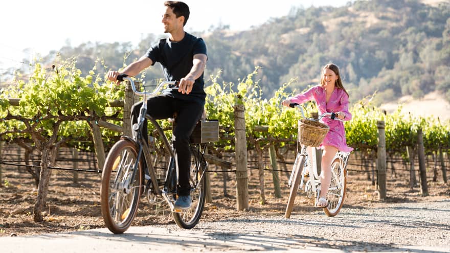 A couple rides bicycles down a dirt path flanked by beautiful vineyards, enjoying the sunshine outdoors.