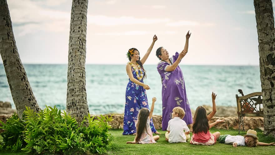 Young children sit on lawn by ocean, watch two Hawaiian women demonstrate dances