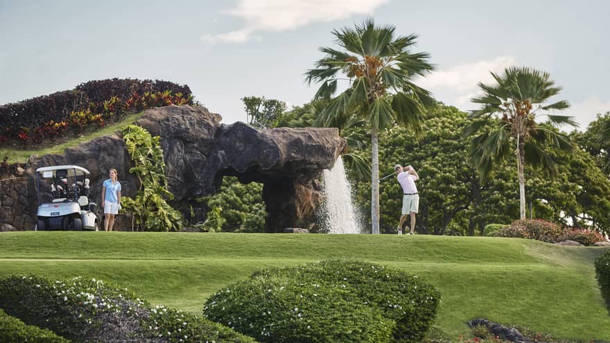 Man swings golf club on course by rock and waterfall, woman stands by cart