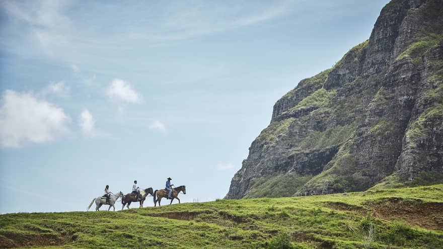 Long side view of three horseback riders on a grassy hill heading toward an imposing verdant cliff under clear blue skies.