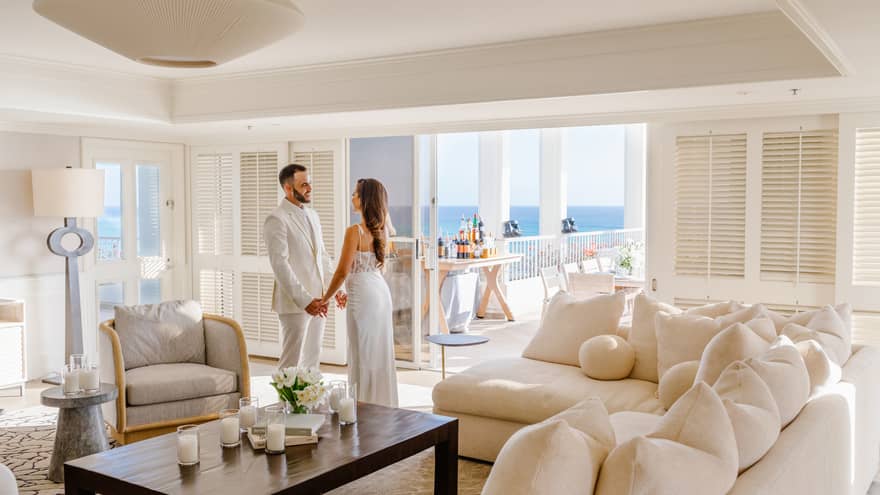 Man and woman stand holding hands in living cream with cream-coloured sofa and walls; opens to ocean-view terrace