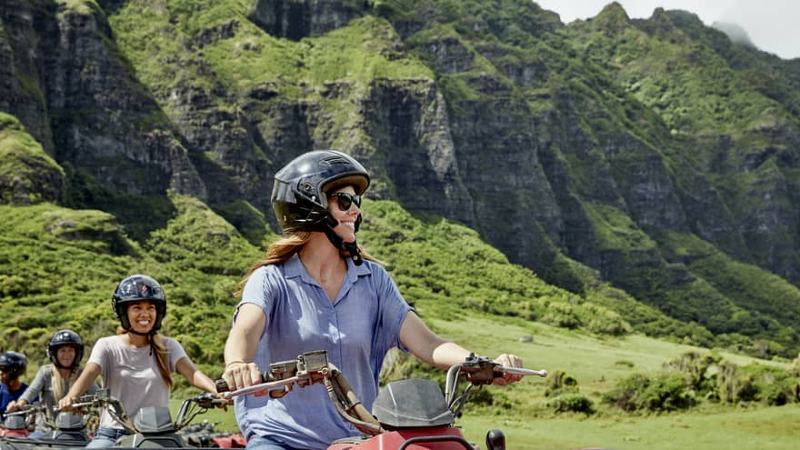 A row of smiling ATV riders wearing black helmets. Behind, a sun-drenched verdant mountain with a mid-layer of craggy rock.