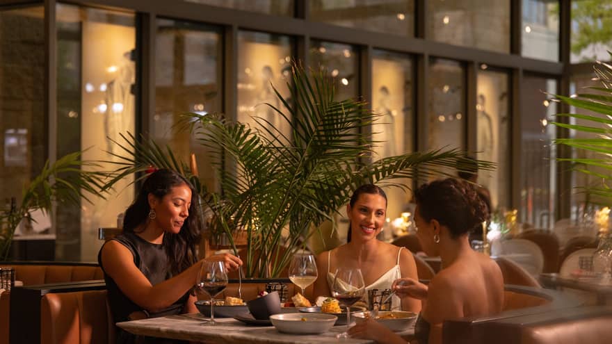 Guests sit at a dining table and smile at one another in a restaurant with floor-to-ceiling windows