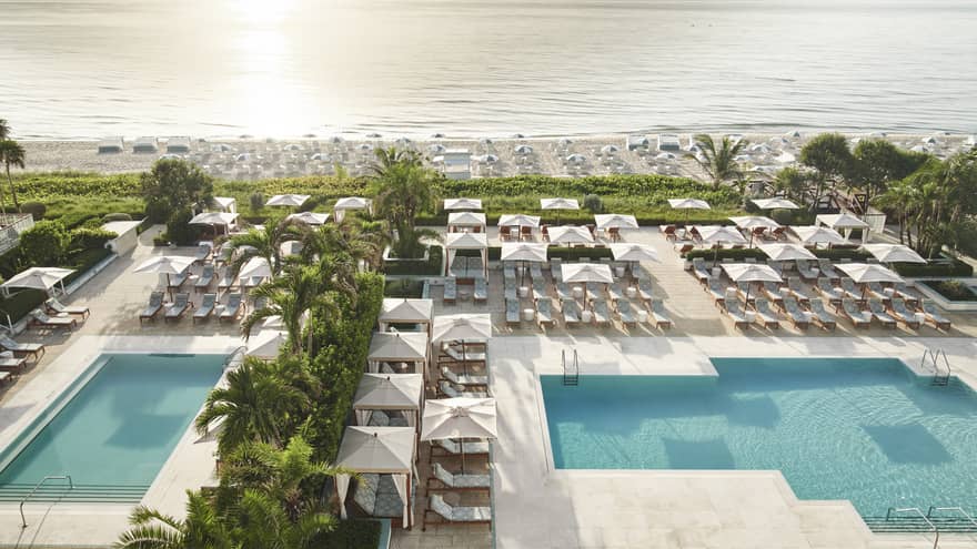 A pool area near a beach with many lounge chairs and umbrellas, surrounded by palm trees.