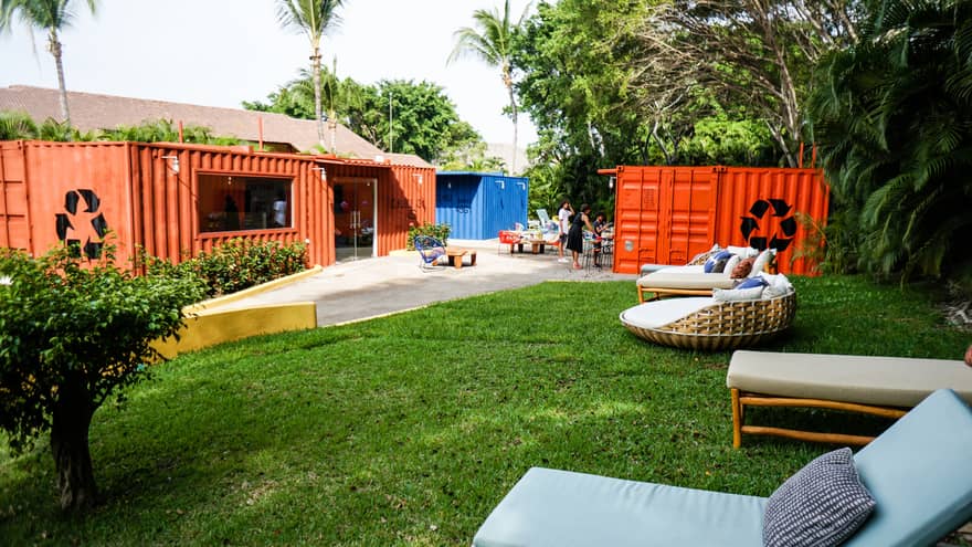 Orange shipping containers turned into rooms for kids and teenagers to play in. There is green grass and chairs outside.