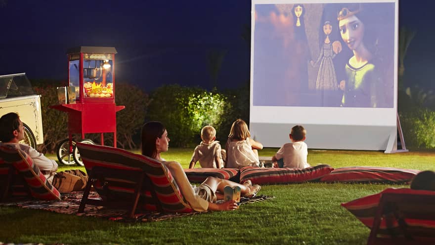 Children, parents lounge on pillows on grass in front of large outdoor movie screen at night