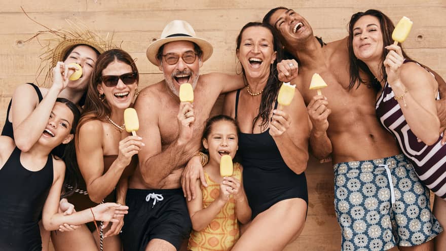 A group of adults and children in bathing suits hold yellow popsicles and smile at the camera.