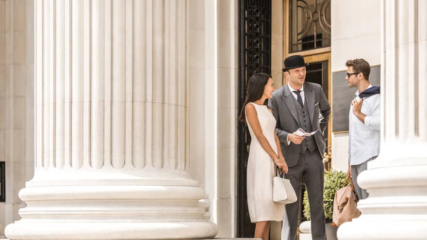 A couple converses with a Concierge on sunny steps beneath a large white pillar outside Four Seasons Hotel London at Ten Trinity Square.