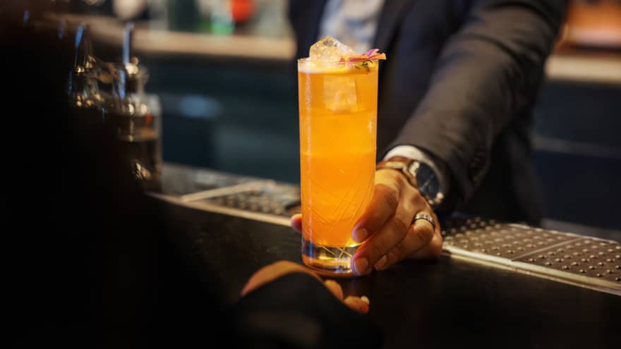 A person sets a bright orange drink in a tall clear glass on a bar counter.