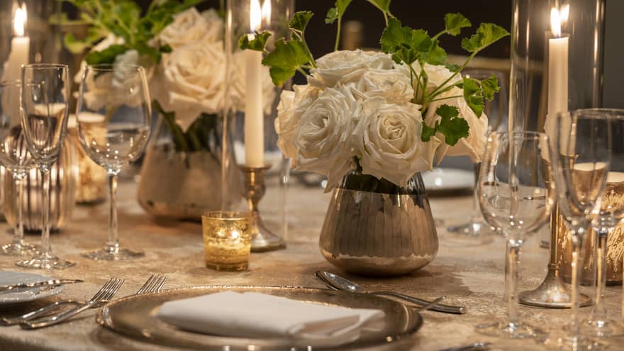 A dining table with white flower center pieces, silver plates, and wine glasses.