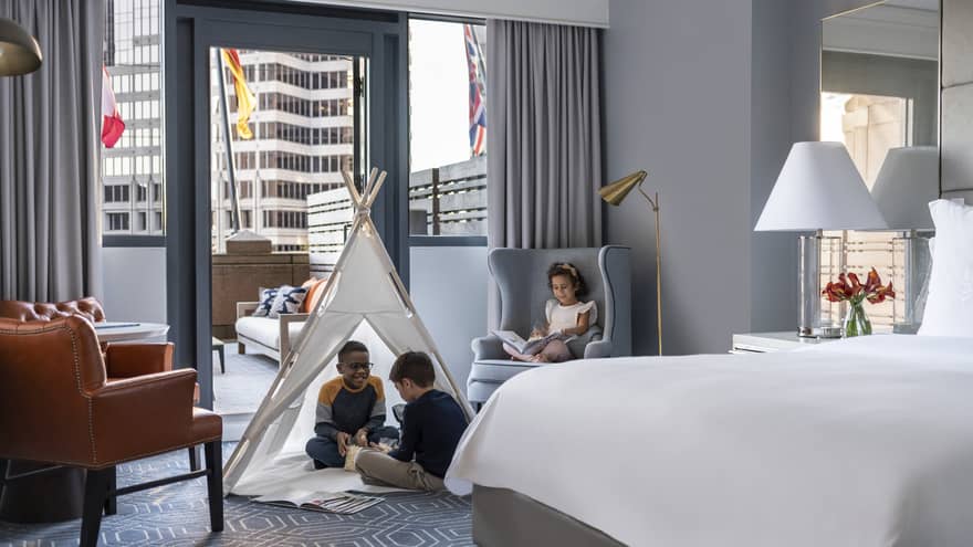 Children sitting in a tent and on a chair in a hotel room.