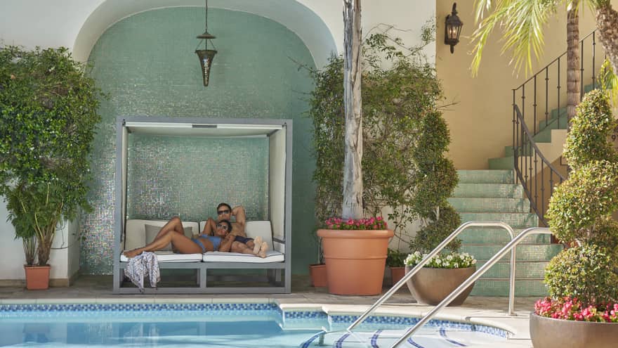 Two people sitting on a lounge chair next to a pool with plants and flowers around it.