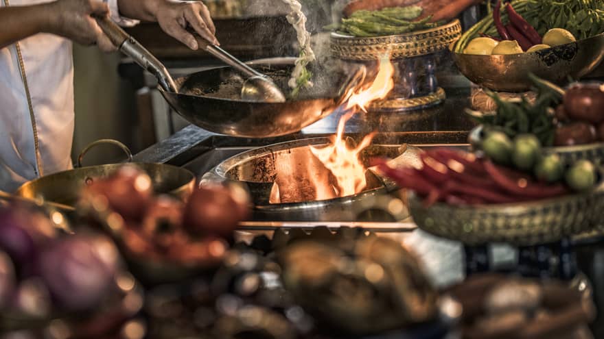Close-up of chef stirring meals in copper pans over stove, flames 