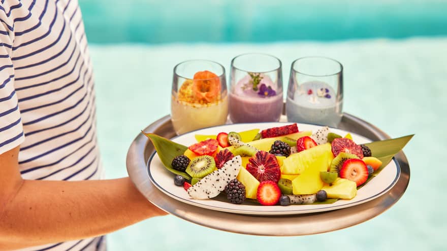 Person carrying tray of smoothies and fruit by pool