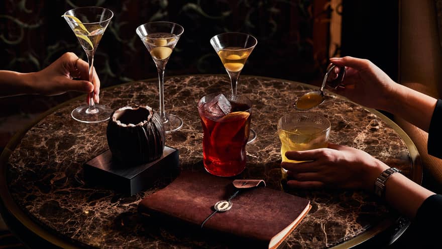 Marble table with assorted cocktails, a brown leather notebook and hands holding martini and whiskey glasses.