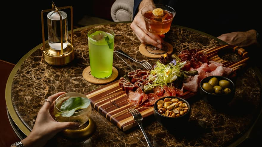 Table with various cocktails and bar snacks on it in dimly lit lounge
