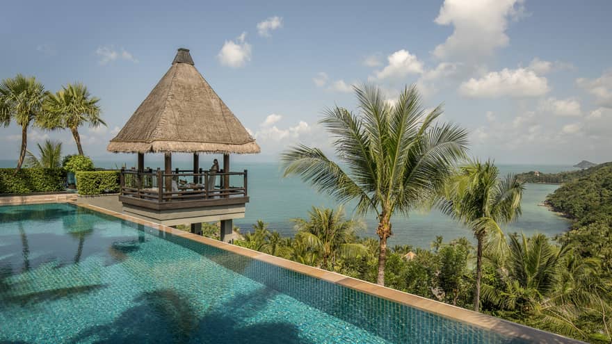 Thatched-roof pavilion at edge of outdoor swimming pool overlooking tropical trees, mountain, ocean