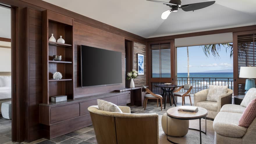 A suite's living room, with off-white sofa, two arm chairs, table, hanging flat-screen TV, walk-out balcony