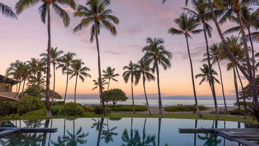 Luxury outdoor resort pool with view of ocean at sunset, surrounded by palm trees, at Four Seasons Resort Hualalai, Hawaii