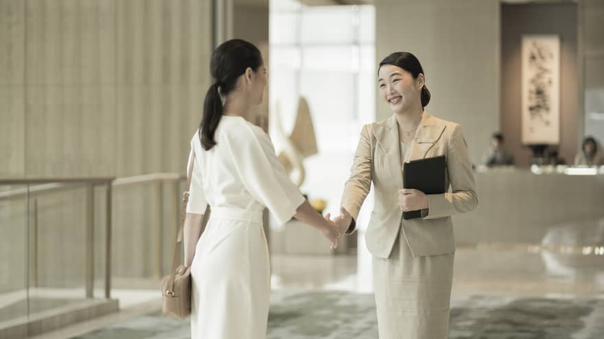 Two women shake hands in a lobby with grey carpeting, in front of a welcome desk.