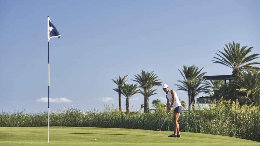 On a bumpy green bordered by reeds and palm trees, a golfer watches the ball's movement toward a hole marked by a blue flag. 
