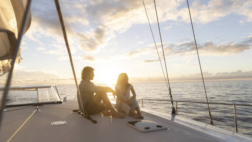 Couple in silhouette sitting barefoot on a boat deck framed by rigging ropes against a bright blue clouded sky at sunset.