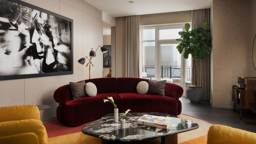 Luxury suite living room with burgundy sofa, coffee table, two yellow armchairs, large black-and-white wall art, potted plant and city-view balcony