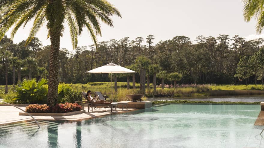 Outdoor swimming pool where woman lounges on patio chair under palm tree 