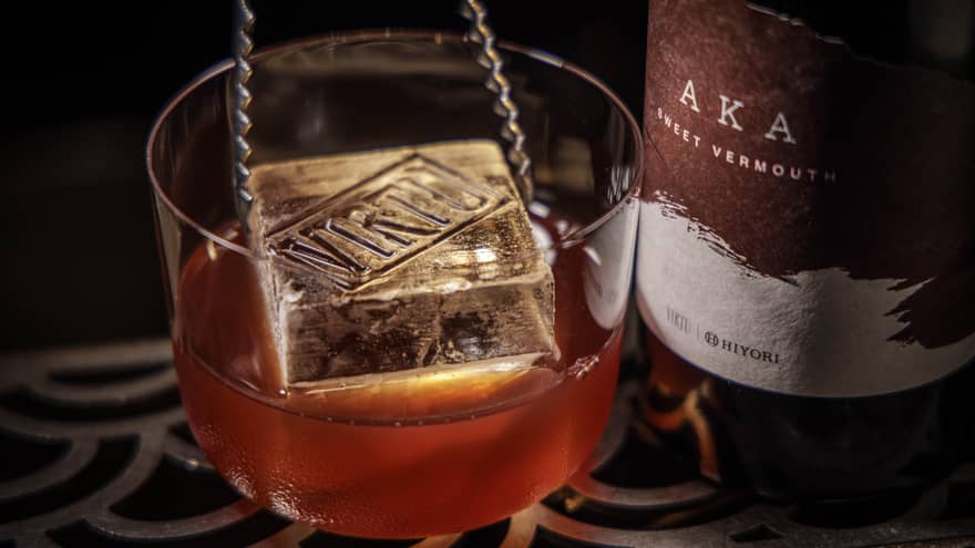 A pair of metal tongs lowers a large square ice cube into a chilled cocktail next to a bottle of Vermouth