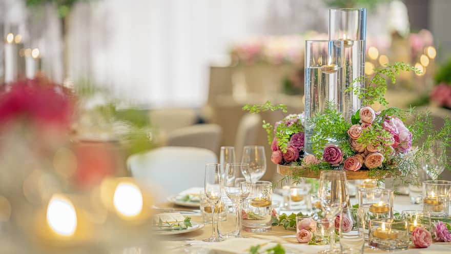 A wedding table setting featuring soft flickering candles in long glass holders, pink roses centerpiece and champagne glasses