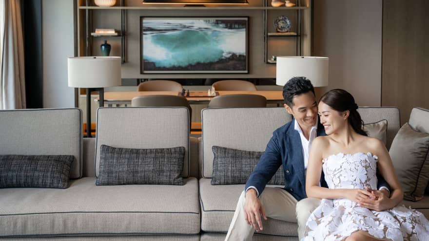 Smiling couple seated on a light gray sofa in a modern living room with decorative shelves and a large ocean artwork in the background.