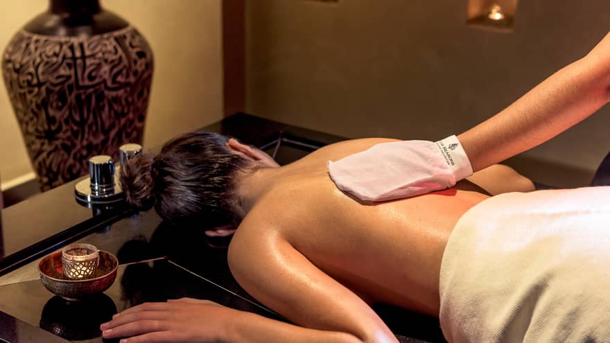 Woman lies face-down on spa table while hand with white mitten massages her back