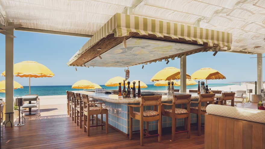 An open outdoor dining area, partially shaded by an off-white translucent awning, with wood deck flooring, seating, and bar counters and tables, some of which have yellow umbrellas for shade. The beach, the ocean and a clear blue sky are in the background.