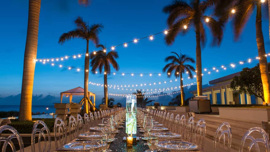 Long outdoor banquet dining table under string lights, palm trees at dusk 