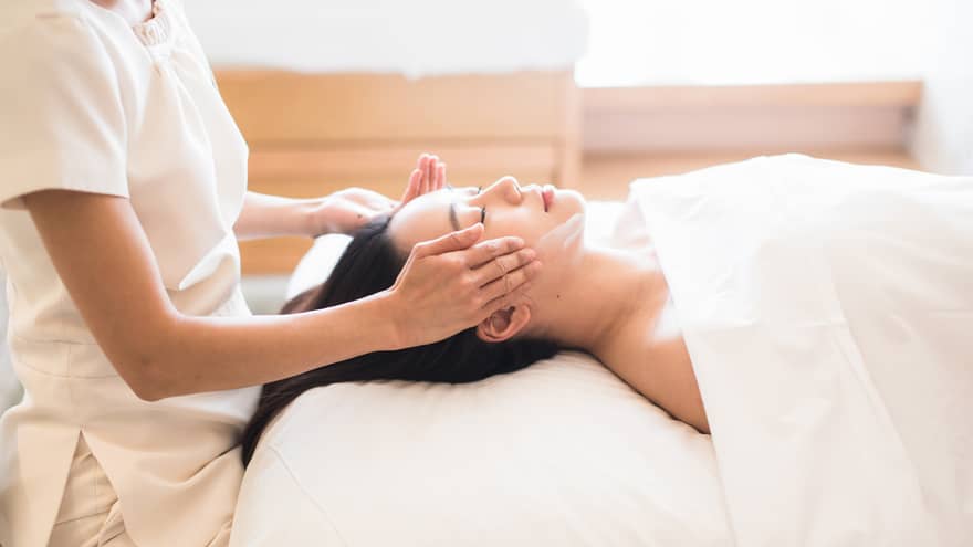 Woman with eyes closed rests head on white pillow as spa attendant rubs cream along her cheekbones