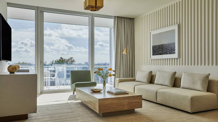 Low beige sofa, wood table in bright seating area by glass balcony doors. This beautiful room is part of the private residences at Four Seasons Hotel at the Surf Club, Surfside, where luxury vacation rentals in Miami are luxuriously situated.