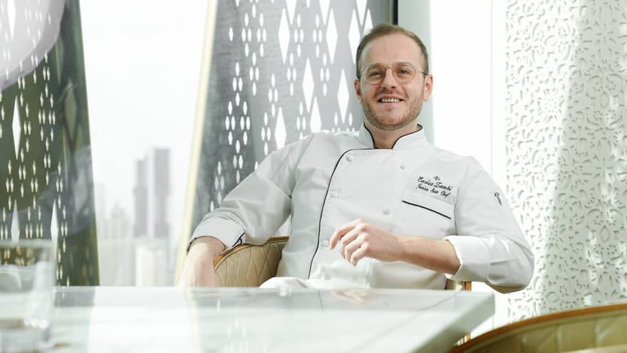 Chef with short light-brown hair, slight beard and wearing white chef's jacket sits back in a cream-coloured club chair at a restaurant table with a city skyline in the background