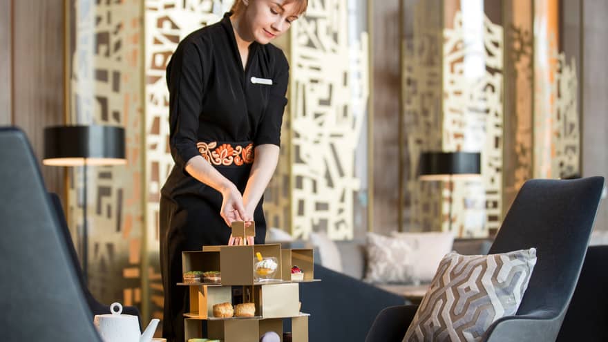 Server holds geometric gold tray with Afternoon Tea small gourmet desserts on lounge table near modern grey chair, pillow