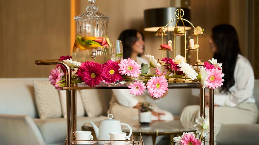 Bar cart decorated with pink Gerber daises carries a gold tray of pastries, a white tea pot and two mugs, and a large glass carafe of fruit-infused water