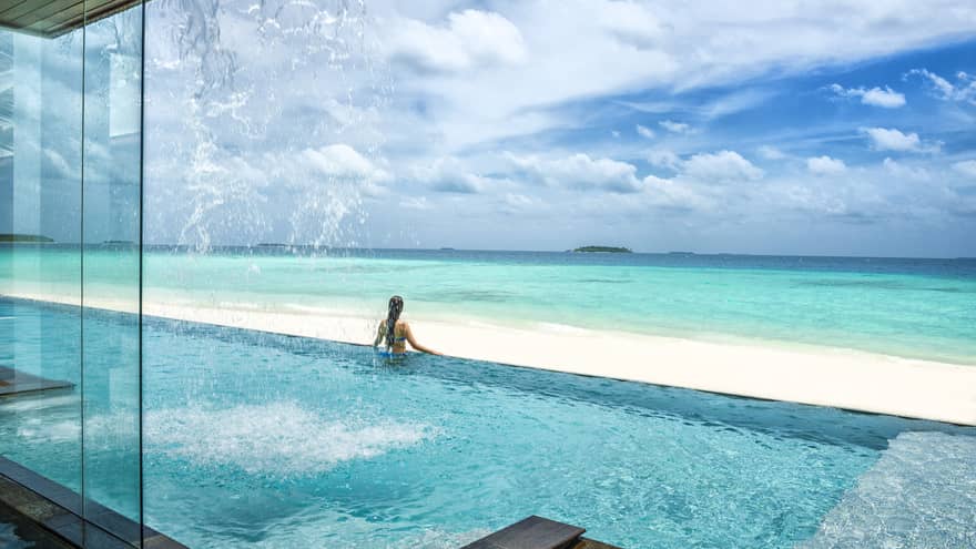 Woman stands at edge of infinity swimming pool looking out over white sand beach, blue ocean