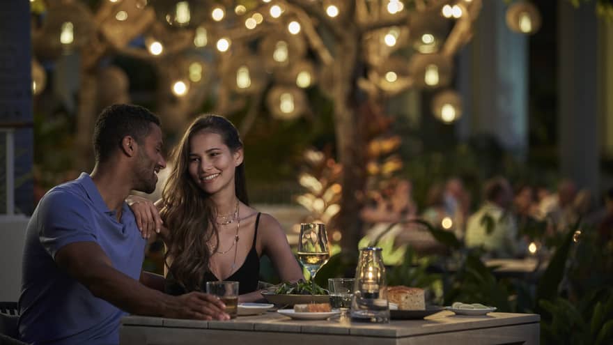 A couple sits together at a dining table beneath a tree lit with twinkling string lights