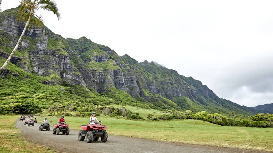 A row of ATV riders ride toward the foreground, leaving behind a verdant mountain with craggy rock rising into misty clouds.