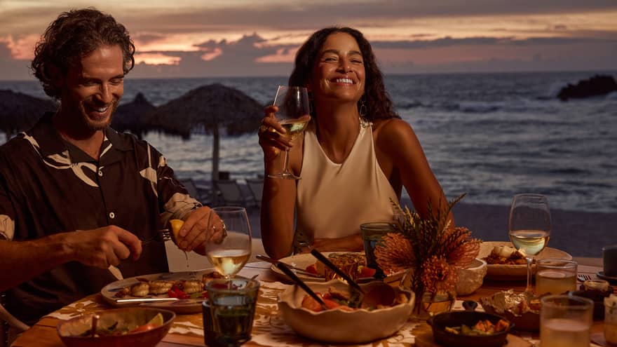 Man and woman laugh while dining at outdoor table filled with food and wine, next to beach and ocean