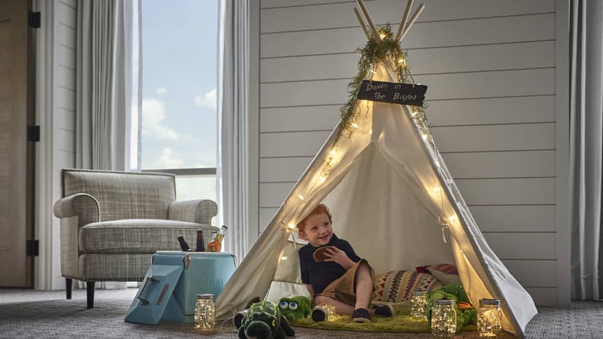 In a small tepee, a smiling child holds a cookie and is surrounded by pillows, stuffed animals and jars of twinkling lights.