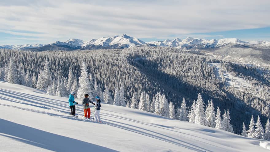Three guests on skis pause on the ski hill, snow capped trees and mountains in distance 