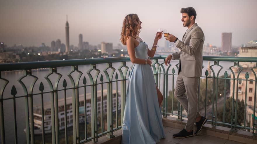 A couple in formal attire clink champagne glasses on a rooftop terrace, view of the river and city skyline beyond. 