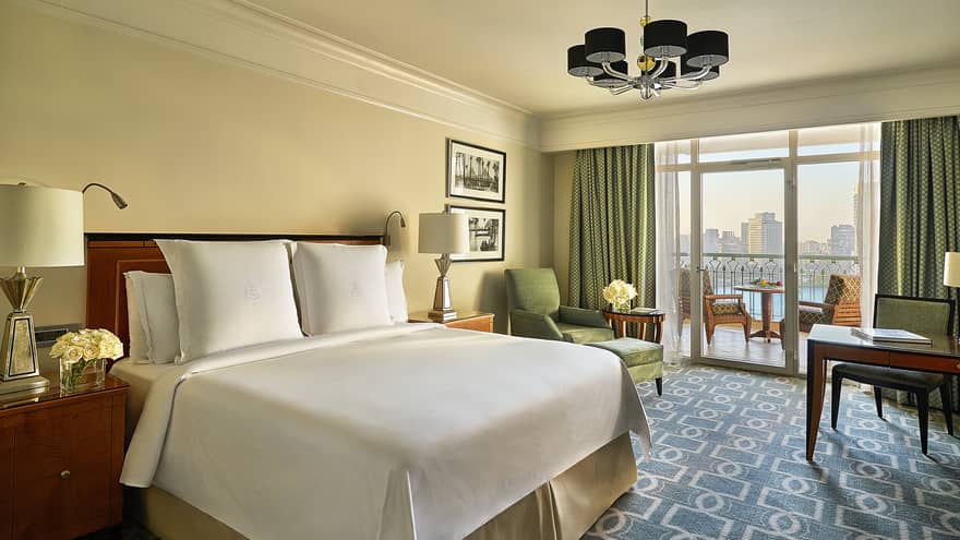 Nile View Room with white bed, blue-and-white carpet, green armchair and curtains leading to balcony