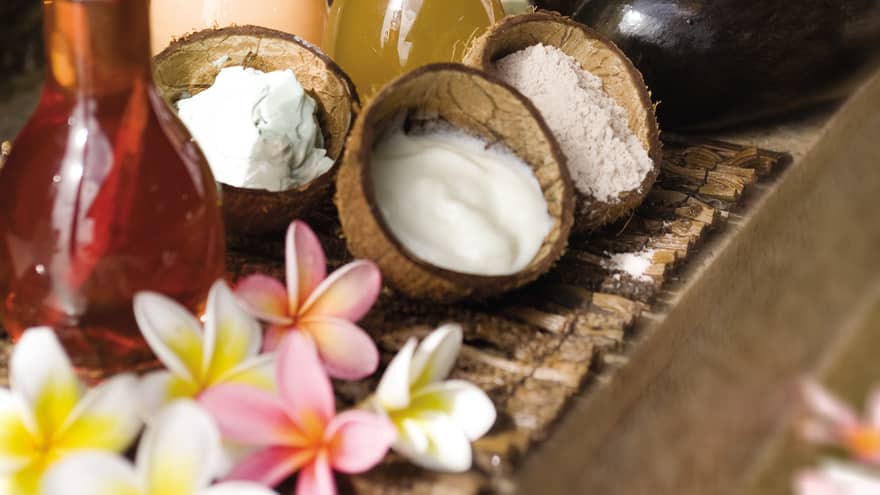 Three open coconut shells filled with white creams and lotions, glass bottles with infused oils, flowers