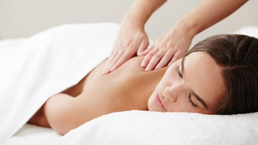 Woman with eyes closed lies under white sheet on table as hands massage her shoulders